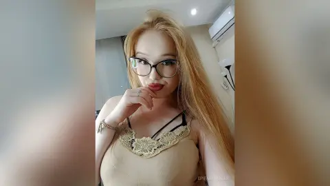 Blonde woman with glasses in a beige lace camisole posing in a bright indoor setting.