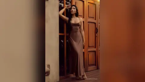 Elegant woman in a sleek brown satin slip dress leaning against a wooden door in soft, moody lighting.