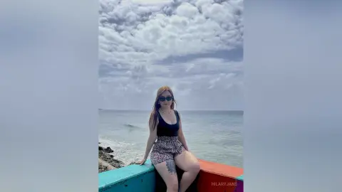 Young woman in black tank top and floral shorts relaxing by the ocean under a cloudy sky.