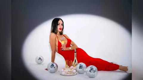 Elegant woman in a red evening gown holding champagne near disco balls and a cake in a celebratory studio setting.