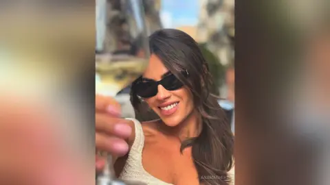 AnaMathers smiles while wearing black sunglasses and a white knit top, holding a glass of white wine outdoors.