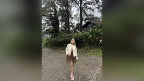 Blonde woman in a white cardigan and brown tulle skirt standing on a forest path under tall trees.