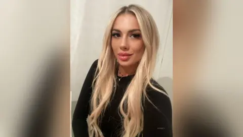 Blonde woman in a black long-sleeve top posing with a glamorous makeup look in a bright indoor setting.