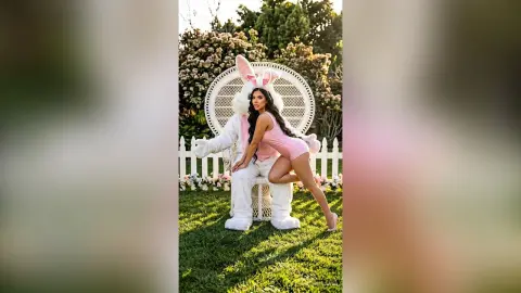 Woman in a pink bodysuit and bunny ears posing in a garden setting with a whimsical, playful mood.
