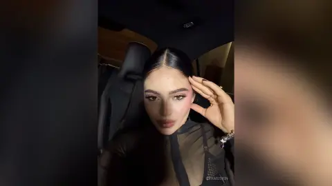 Young woman with sleek dark hair and glam makeup wearing a black sheer top inside a car at night.