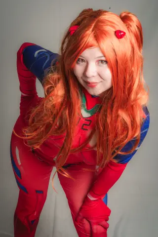 Cosplayer in a red and blue superhero bodysuit with long wavy orange hair posing against a white background.