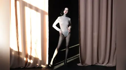 Elegant woman in a white long-sleeve bodysuit and black tights posing between beige curtains in dramatic lighting.