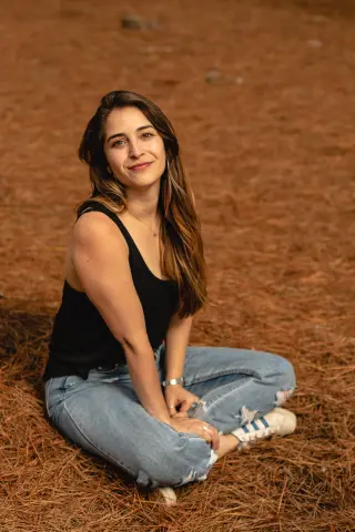 Smiling young woman in a black tank top and blue jeans sitting on dry grass in a warm outdoor setting.