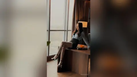 Elegant woman in a grey long-sleeve top and black skirt sitting by a high-rise window with a city view.