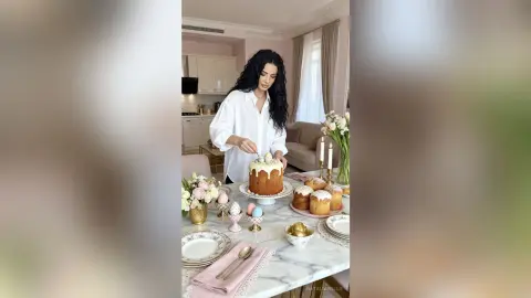 Woman in white shirt serving traditional Easter bread and decorated eggs on a marble dining table.