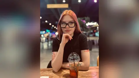 Red-haired woman in a black t-shirt sitting at a cafe table with an iced drink in a moody indoor setting.