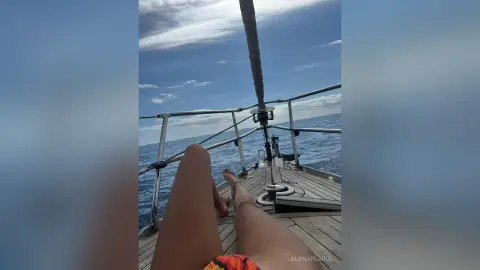 AlanaPearce relaxing on a wooden boat deck under a blue sky, showing her tanned legs and feet while wearing orange and red swimwear.