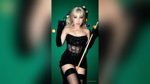 Blonde woman in a black lace corset and mini dress holding a pool cue against a green background.