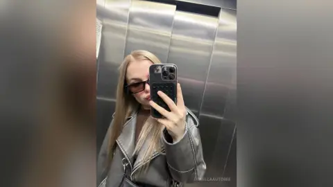 Stylish blonde woman in a grey leather jacket and sunglasses taking a mirror selfie in a modern elevator.