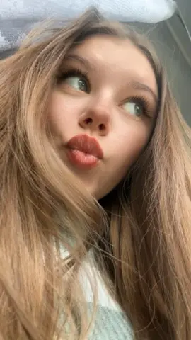 Young woman with long blonde hair and red lipstick making a playful pouty face in a bright setting.