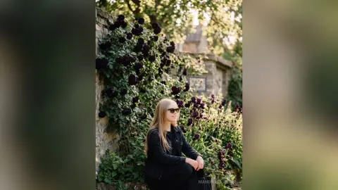 Young woman in a black denim jacket wearing sunglasses sitting in a lush summer garden with dark flowers.