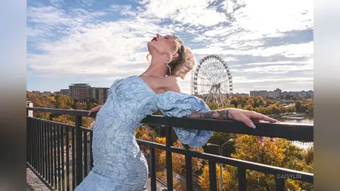 DaisyLin leans back against a black metal railing wearing a light blue off-the-shoulder lace dress with her blonde hair styled up.