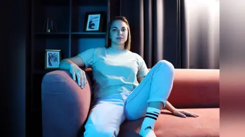 Young woman in a light blue t-shirt and jeans relaxing on a pink sofa in a moody, dimly lit room.
