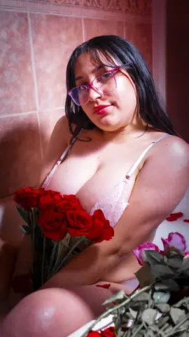 Young woman in pink lace lingerie holding red roses in a romantic, warm-lit bathroom setting.