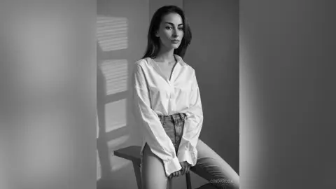 Elegant woman in a white button-down shirt and jeans sitting in a moody, sunlit indoor setting.