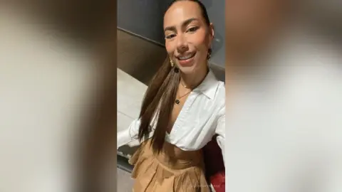 VictoriaFontaine smiles, wearing a white button-down shirt and tan skirt, with her long brown hair swept to one side.
