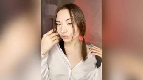Young woman in a white button-down shirt and silver necklace posing with a playful expression in soft indoor lighting.