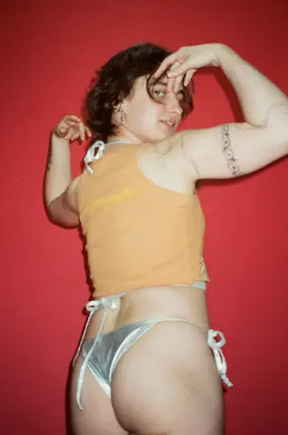Young woman in an orange crop top and silver bikini posing against a vibrant red background.