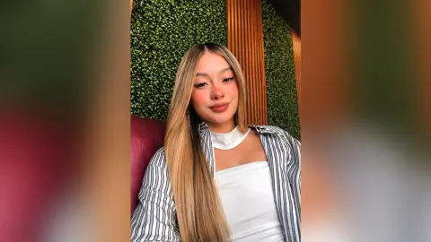 Smiling young woman with long blonde hair wearing a white top and striped blazer in a modern green lounge.