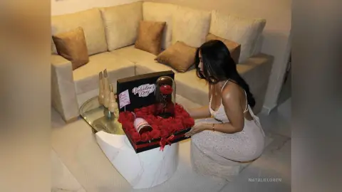 Woman in a white sequin dress sitting by a sofa with a romantic Valentine's Day rose gift box.