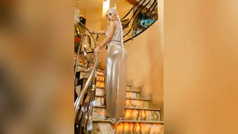 Anastasia stands on a stone staircase wearing a long, shimmering gold gown that clings to her curves.