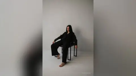 Elegant woman in a black oversized suit sitting on a chair against a minimalist white studio background.