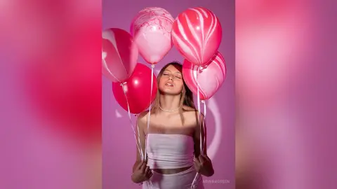 ArinnaGreen poses with pink and white swirl balloons, wearing a white strapless top against a pink background.