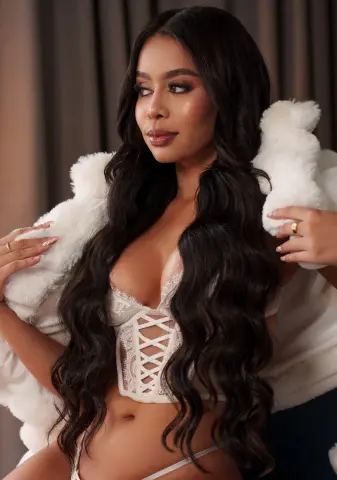 Glamorous woman with long wavy hair wearing a white lace corset and fluffy faux fur coat in a soft studio setting.