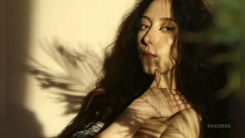 Moody portrait of a woman with dark wavy hair and winged eyeliner in warm sunlight and palm shadows.