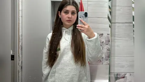 Young woman with long brown hair wearing a cozy white hoodie taking a mirror selfie in a bright room.