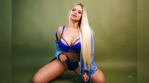 Gia poses in a blue bra and matching panties, wearing an open denim jacket against a green background.