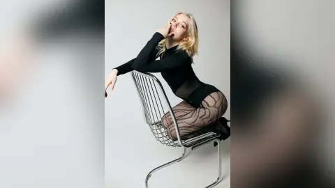 Blonde woman in a black long-sleeve top and patterned tights posing on a silver chair against a white background.