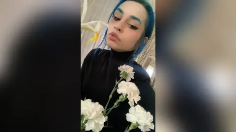 Young woman with blue hair in a black turtleneck holding white carnations in a soft, moody indoor setting.