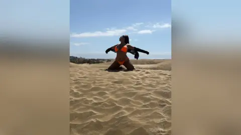 MissyJolie poses in an orange bikini while kneeling on a sand dune under a clear blue sky.