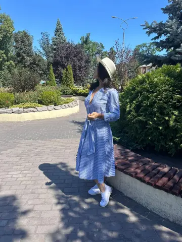 Woman in a blue striped summer dress and straw hat walking on a sunny garden path.