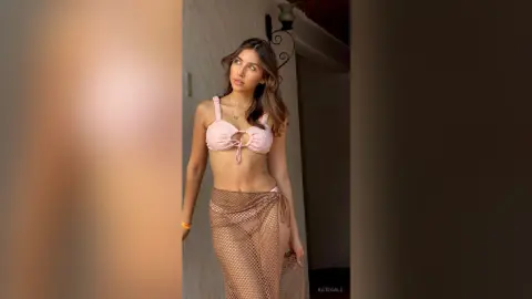 Young woman in a pink bikini top and sheer mesh skirt posing confidently in a warm, sunlit indoor setting.
