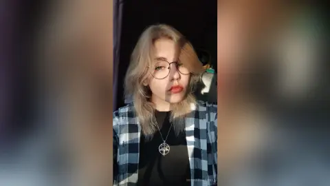 Blonde woman with glasses in a black shirt and plaid flannel shirt posing in soft indoor lighting.