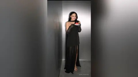 Elegant woman in a black evening gown and gloves holding a small cake in a minimalist studio setting.