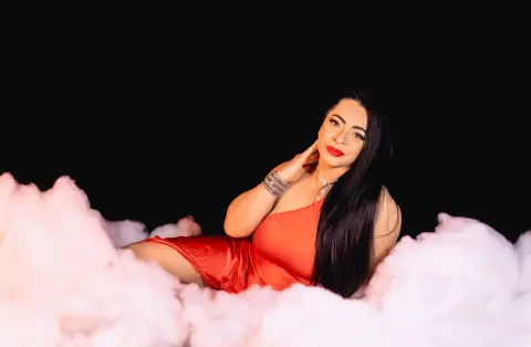 Glamorous woman in a red satin dress sitting amidst soft white clouds against a black background.