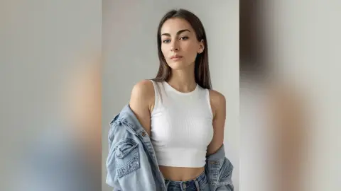 Young woman in a white ribbed tank top and denim jacket posing against a neutral studio background.
