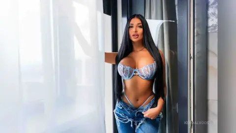 Beautiful brunette woman in a blue lace bra and denim jeans posing by a bright window in a modern room.