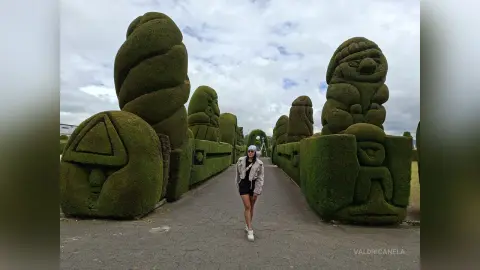 Young woman in a white jacket standing on a path between giant whimsical topiary sculptures under a cloudy sky.