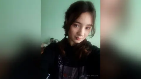 Young woman with long dark hair wearing a black graphic hoodie in a minimalist indoor setting.