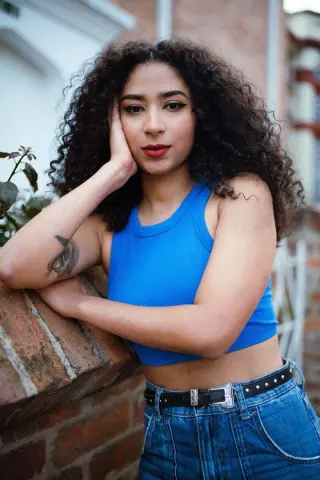 Young woman with curly hair in a blue crop top leaning on a brick wall in a relaxed outdoor setting.