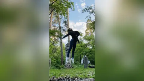 Adventurous woman in dark activewear jumping in a lush green forest under a bright blue sky.
